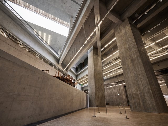 View from the bottom floor looking up through an opening in the floors above. Two concrete pillars hold up the ceiling on our right. On our left, part of an installation artwork consisting of wooden chairs attached to a round table is visible on the level above.